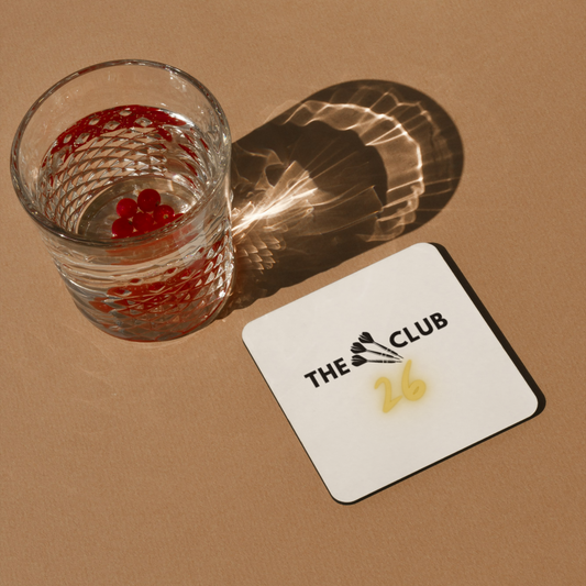 Two glasses with drinks on a brown surface, one with a coaster displaying 'The 26 Club' logo.