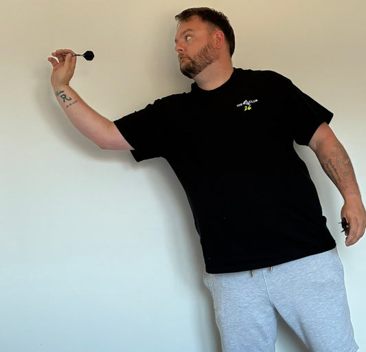 Man in black t-shirt and gray shorts holding a dart against a plain wall