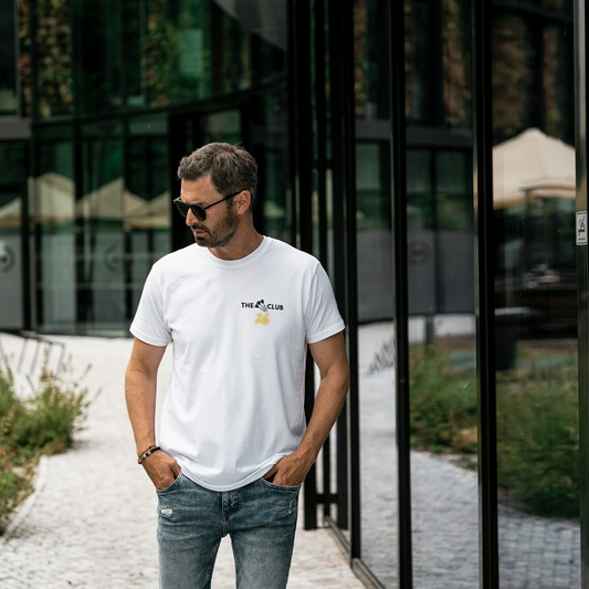 Man wearing a white t-shirt with a The 26 Club logo, standing outdoors near a modern building.