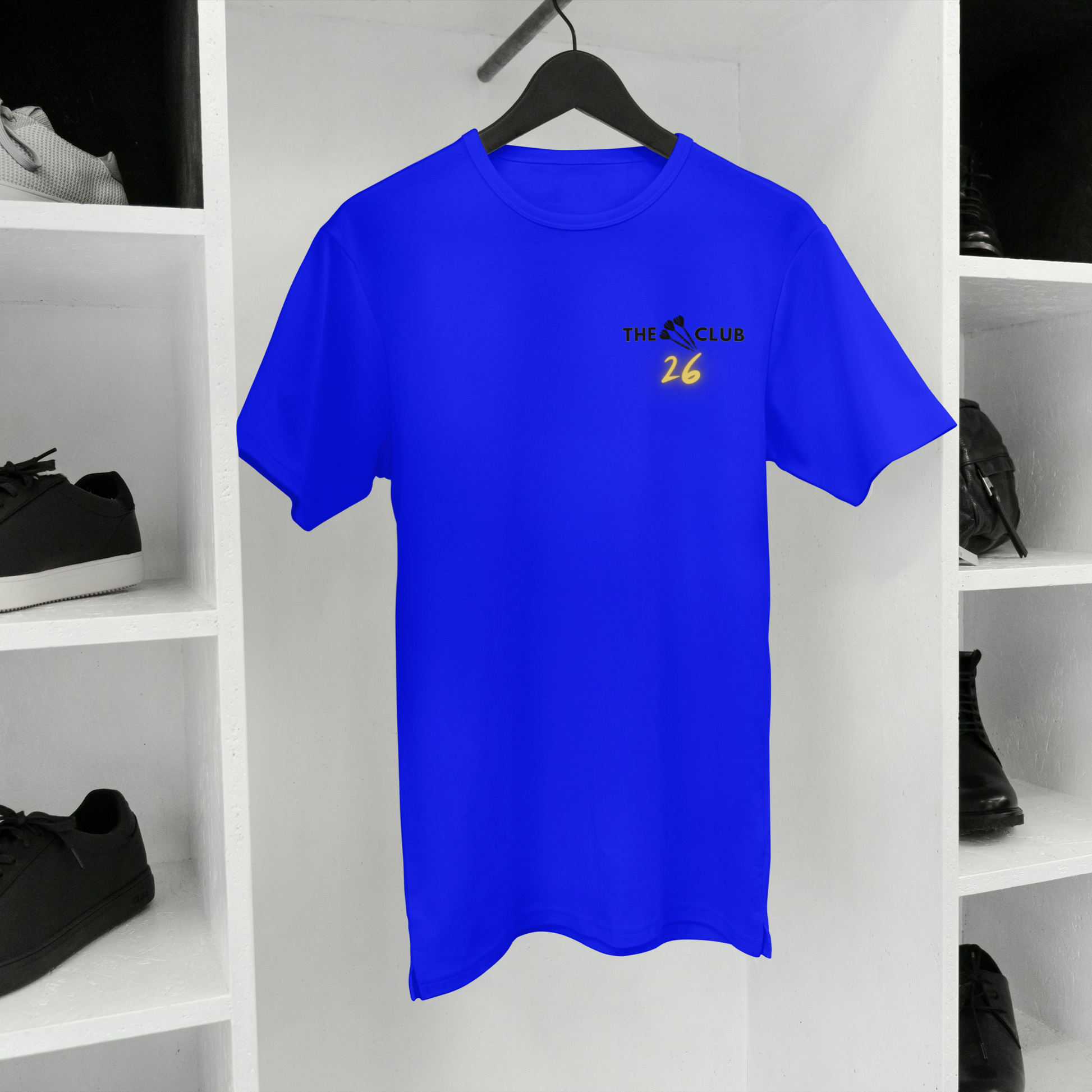 Blue t-shirt with a The 26 Club logo on a hanger against a white shelf background with shoes and clothes.