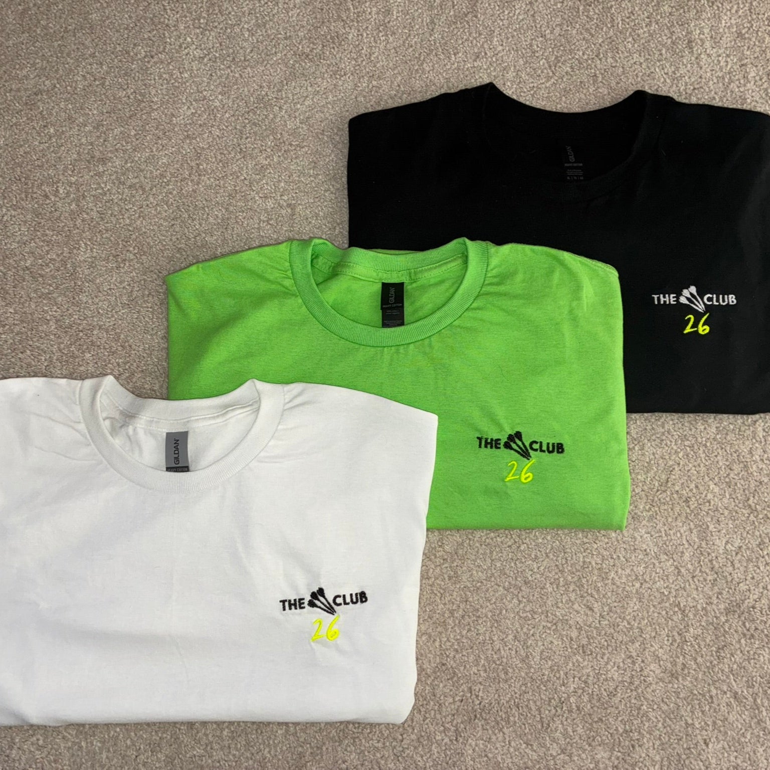 Three folded t-shirts in white, green, and black on a beige carpet.